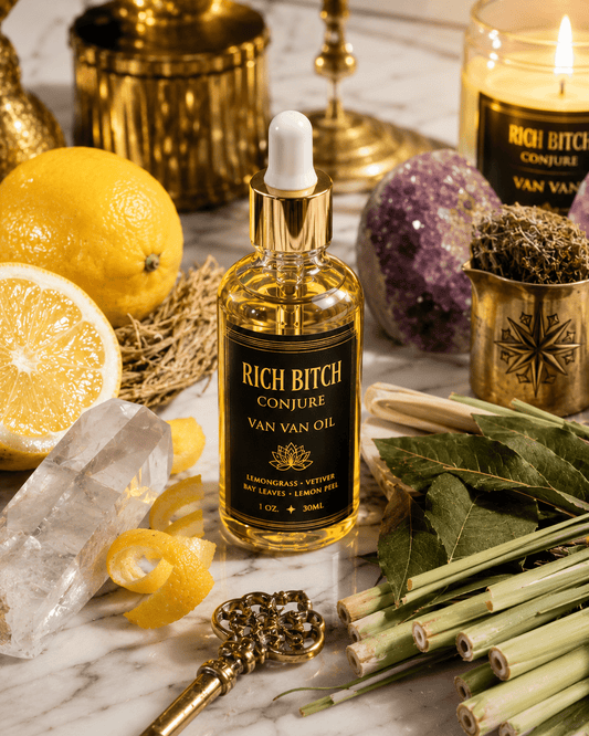 Bottle of 'Rich Bitch Conjure Van Van Oil' surrounded by lemons, crystals, and herbs on a marble surface.